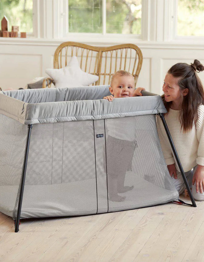 Matka bawiąca się z dzieckiem w łóżeczku turystycznym Baby Bjorn w jasnym, przestronnym pokoju z wiklinowym fotelem i dekoracjami w tle. Wybierz idealne łóżeczko turystyczne dla Twojego dziecka.